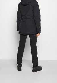 Eine Person in einer schwarzen Kapuzenjacke, schwarzen Hosen und schwarzen Wanderschuhen steht mit dem Rücken zur Kamera vor einem einfarbigen Hintergrund.