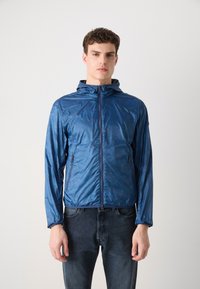 Leichte blaue Jacke mit Kapuze, die eine glatte, glänzende Textur aufweist und über Taschen mit Reißverschluss verfügt. Getragen über dunklen Jeans.