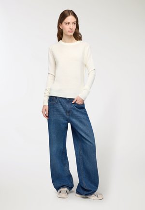 Maglione a maglia crema con maniche lunghe, abbinato a jeans di denim blu oversize. Il modello indossa sneakers chiare. Design semplice e casual.