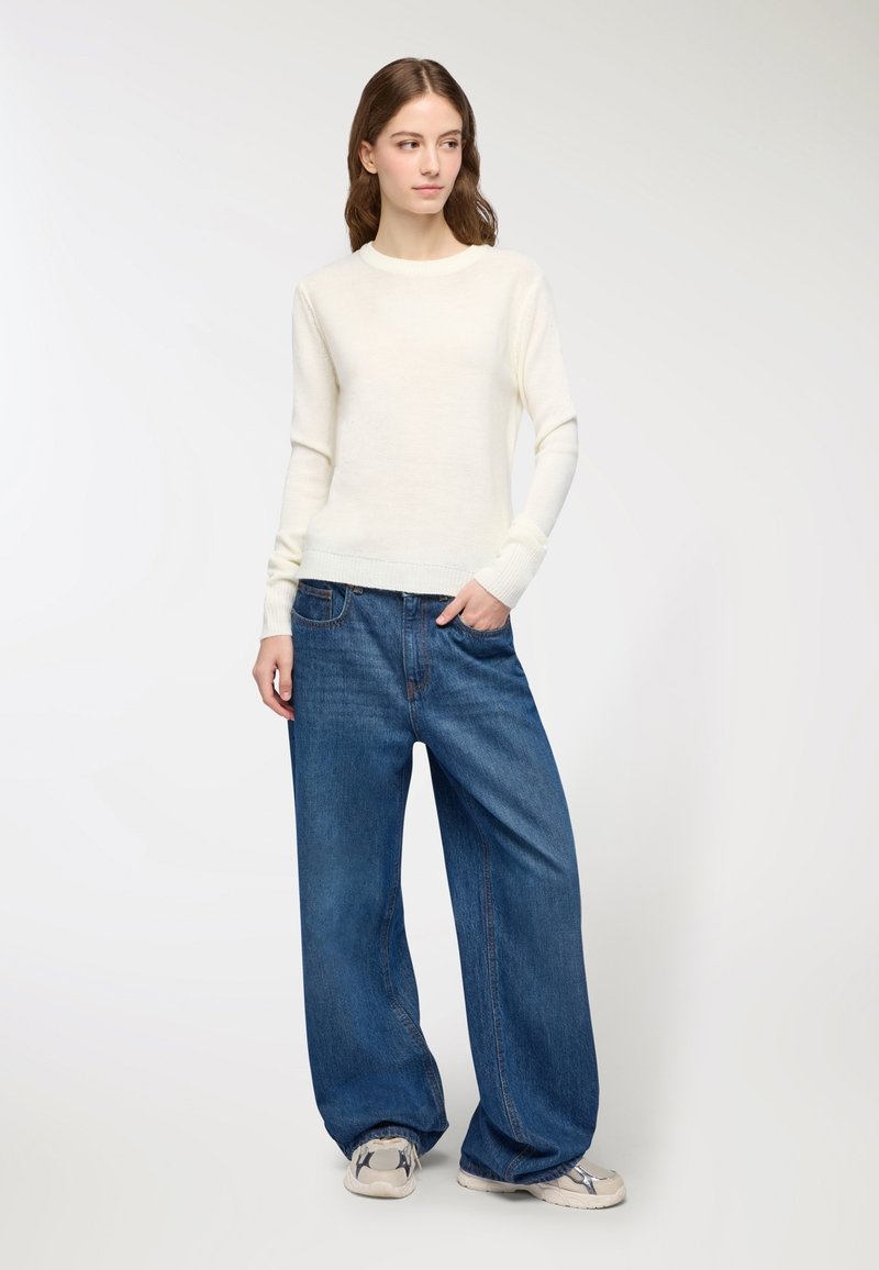 Maglione a maglia crema con maniche lunghe, abbinato a jeans di denim blu oversize. Il modello indossa sneakers chiare. Design semplice e casual.