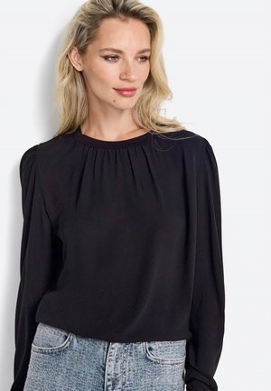 Blouse noire à manches longues avec un col froncé, fabriquée en tissu lisse, associée à un jean en denim bleu clair.