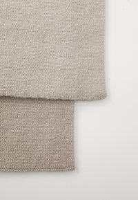 Beige strikket stof med en tekstureret overflade. To overlappende lag viser en lysere øverste sektion og en lidt mørkere bund, med færdige kanter.