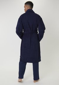 Mann, der barfuß steht, mit dem Rücken zugewandt, einen marineblauen Bademantel und passende Hosen trägt, vor einem schlichten Hintergrund.