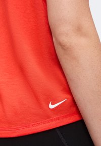 Άτομο που φοράει κόκκινη μπλούζα Nike με ορατό λευκό λογότυπο Nike κοντά στο στρίφωμα, το χέρι ακουμπάει στο πλάι, μαύρο παντελόνι εν μέρει ορατό.
