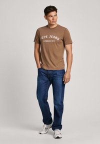 Ruda medvilninė marškinė su balta "Pepe Jeans" užrašu, derinama su tamsiai mėlynomis džinsinėmis kelnėmis. Modelis vilki baltus sportbačius, stovi neutraliame fone.