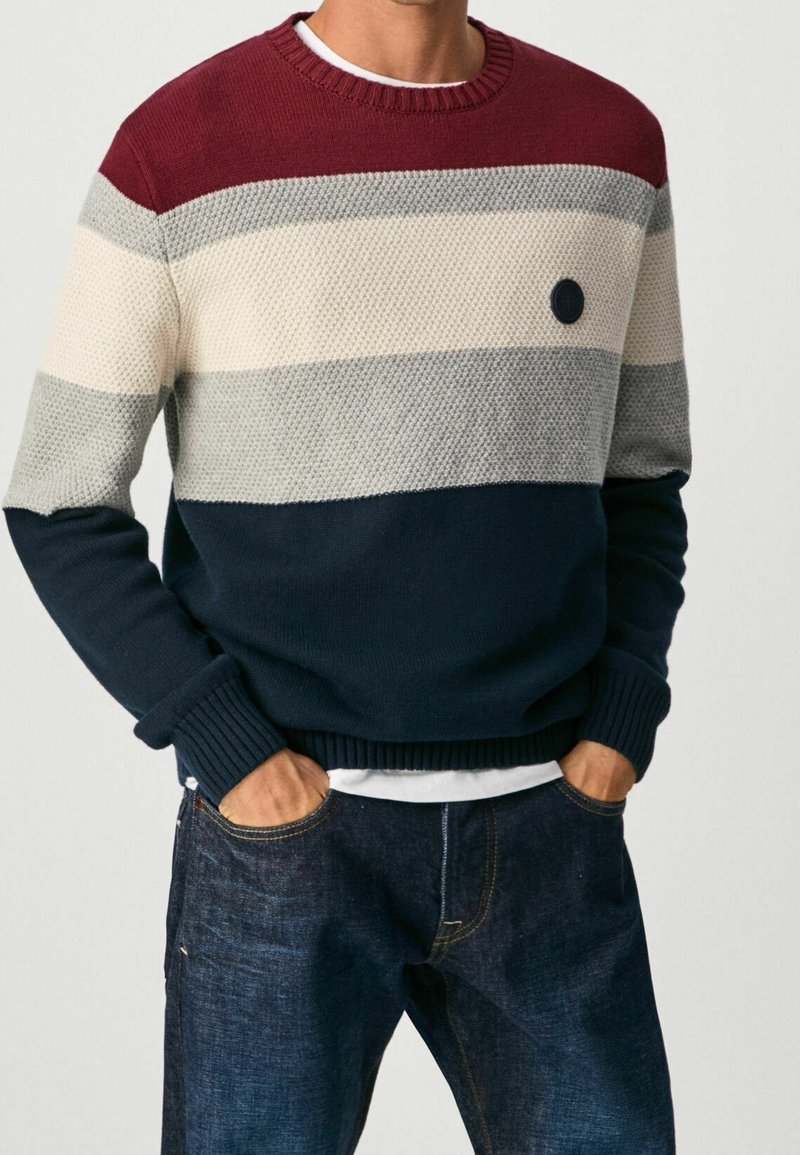 Maglione lavorato a maglia con strisce orizzontali color bordeaux, grigio, beige e blu navy. Presenta un collo rotondo, polsini a coste e una toppa circolare blu navy sul petto.