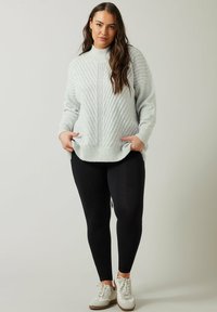 Lichtgrijze gebreide trui met een hoge hals, geribbelde textuur en zijzakken, gecombineerd met zwarte leggings en witte sneakers.