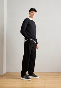 Fekete pulóver egy fehér galléros ing felett, fekete széles szárú nadrág és fekete sportcipő fehér talppal. A modell egy semleges háttér előtt áll.