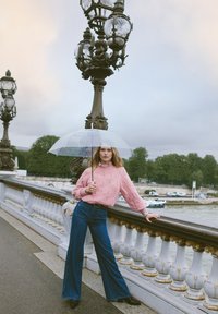 Suéter de punto rosa con un patrón de diamantes, pantalones de mezclilla acampanados de cintura alta y un paraguas transparente. Entorno urbano con una farola ornamentada.