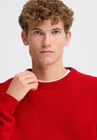 Czerwony dzianinowy sweter z prążkowanym okrągłym dekoltem, teksturowaną powierzchnią i długimi rękawami. Nosi się go na białą koszulkę, w luźnym fasonie.