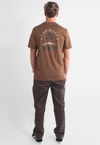 T-shirt en coton marron avec un motif imprimé représentant des palmiers et le mot "SANDWAVES". Associé à un pantalon marron foncé et des baskets noires.
