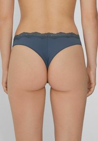 Femme portant un string en dentelle gris foncé, vue de dos sur un fond clair uni.