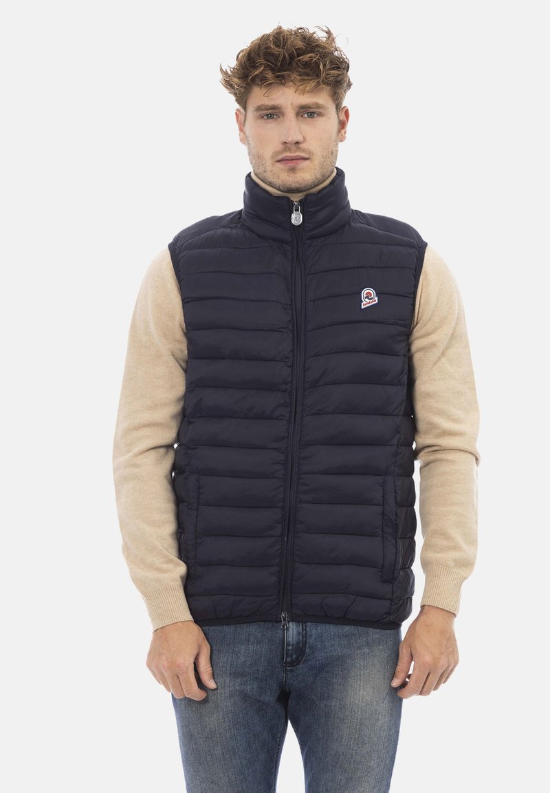 Gilet imbottito blu navy con colletto alto e cerniera frontale, abbinato a un top beige a maniche lunghe. Presenta una patch con il logo sul petto sinistro.