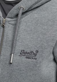 Felpa grigia con zip e cordoncino, con logo "SuperDry" ricamato e caratteri giapponesi sul petto.
