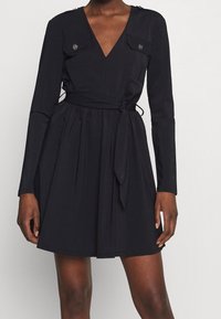 Robe noire avec un décolleté en V profond, des manches longues, deux poches poitrine avec des accents de boutons, et une taille froncée avec une ceinture à nouer. Texture lisse.