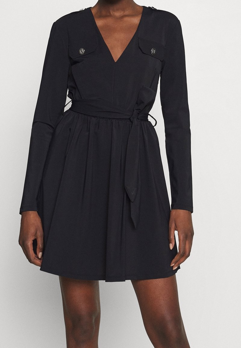 Robe noire avec un décolleté en V profond, des manches longues, deux poches poitrine avec des accents de boutons, et une taille froncée avec une ceinture à nouer. Texture lisse.