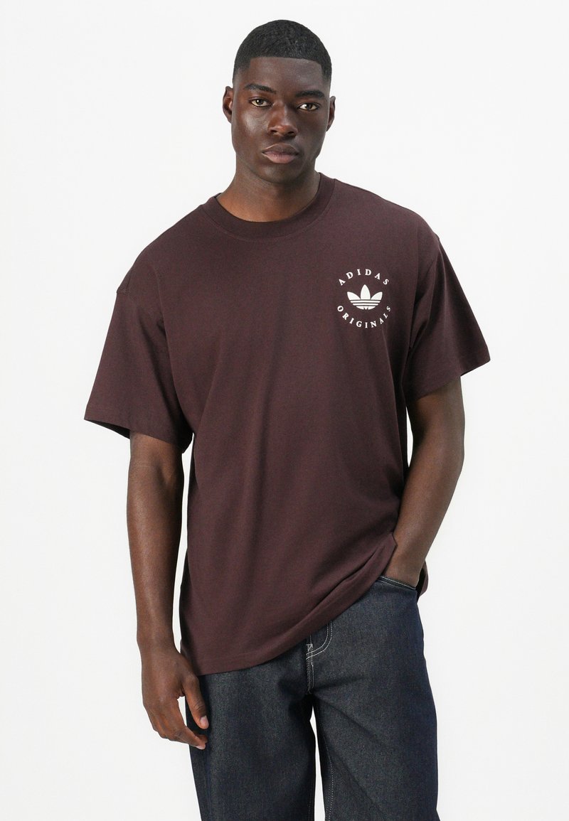 Maglietta in cotone marrone con maniche corte, vestibilità ampia, con logo bianco Adidas sul petto. Abbinata a jeans in denim scuri.