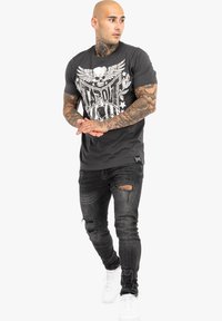 Graphitfarbenes T-Shirt mit einem weißen Graf print, das einen Schädel und Sterne zeigt. Kombiniert mit ausgewaschenen schwarzen Jeans und weißen Sneakers.