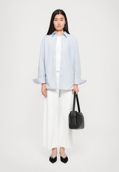 Camicia azzurro chiaro con maniche lunghe, abbinata a pantaloni bianchi a gamba wide leg e ballerine nere a punta. Tiene in mano una borsa nera.