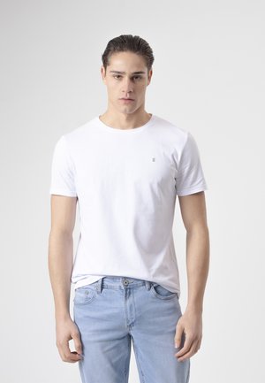 Joven con el pelo peinado hacia atrás, que lleva una camiseta blanca lisa y vaqueros azul claro, de pie frente a un fondo gris.