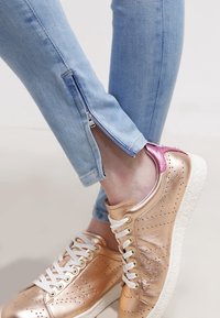 Jeans skinny azul claro con cremalleras laterales, combinados con deportivas metálicas en oro rosa con cordones blancos y una suela texturizada.