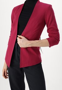 Elegant vínový sako s šálovým límcem, vyrobené z hladké látky. Dlouhé rukávy a otvorený design. Nosené přes černý rolák.