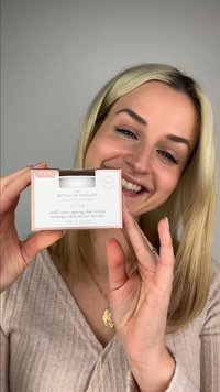 Donna con capelli biondi che sorride, tenendo una confezione di The Ritual of Namaste Glow crema giorno anti-età ricaricabile davanti a uno sfondo neutro.