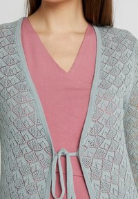 Cardigan tricoté bleu clair avec un motif de dentelle en losanges, présentant une fermeture à nouer à la taille, porté par-dessus un haut rose à col en V.