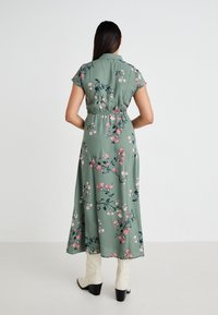 Robe florale verte avec des manches courtes, taille cintrée et longueur cheville. Présente des motifs de fleurs roses et blanches, ainsi qu'une texture de tissu douce.