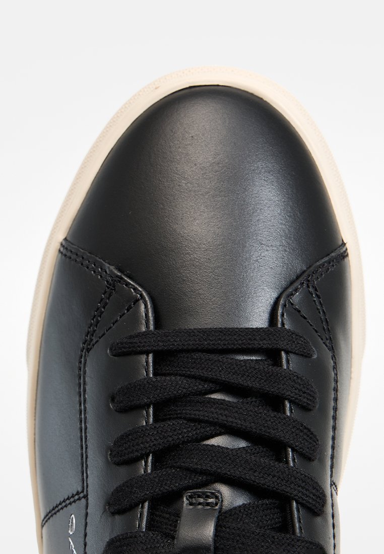 Sneaker in pelle nera con punta tondeggiante, caratterizzata da lacci neri e una suola in gomma color crema. Texture liscia con dettagli di cucitura lungo i lati.