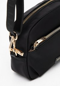 Černá nylonová crossbody taška se zlatými zipy, strukturovaným tvarem a odnímatelným popruhem. Detail loga na přední straně. Hladká textura.