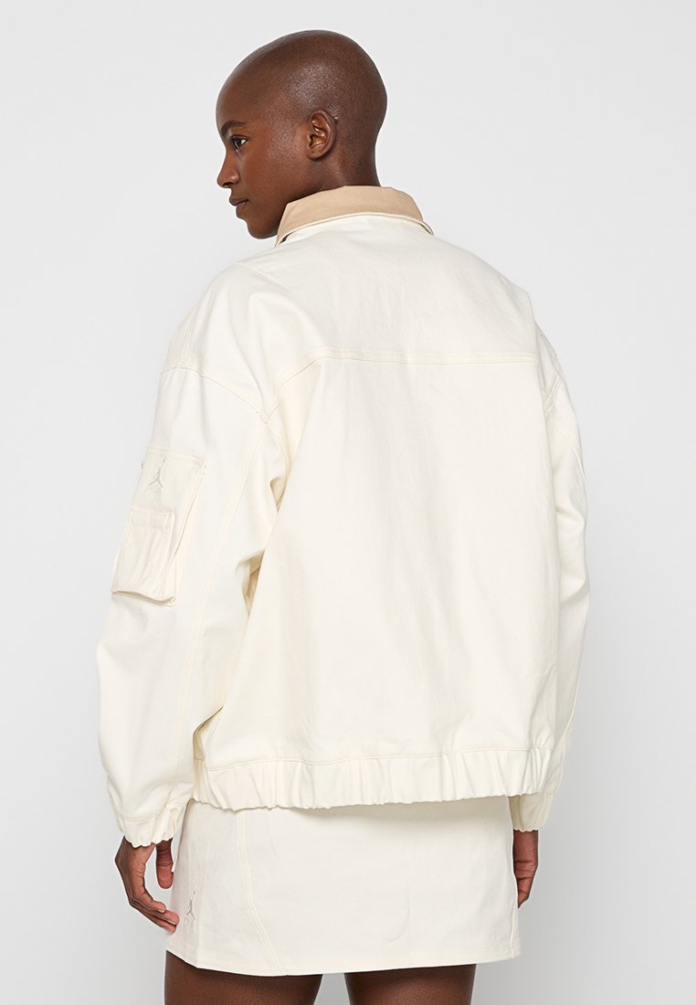Chaqueta de gran tamaño en color crema claro con cuello tipo polo, dobladillo elástico y bolsillo de parche en la manga izquierda. Tela suave con costuras mínimas.