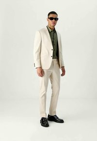 Man wearing cream suit, olive green shirt, black sunglasses, and black dress shoes standing against plain light background.