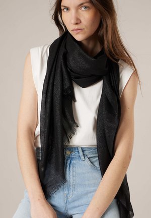 Femme portant un haut blanc sans manches, un jean bleu clair, et une longue écharpe noire à franges drapée autour de son cou et de ses épaules.