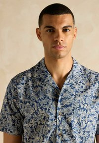 Košela s krátkym rukávom vyrobená z námorníckej látky s béžovým kvetinovým vzorom, s gombíkovou légou a golierom.