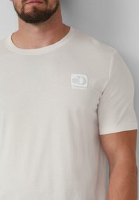 Lichtbeige katoenen t-shirt met korte mouwen, voorzien van een wit logopatch met een wereldbolontwerp op de linkerborst.