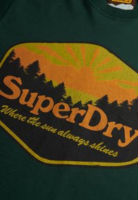 Groen t-shirt met een oranje en gele grafisch ontwerp van bergen, bomen en zonnestralen. Tekst luidt "SuperDry" en "Waar de zon altijd schijnt".