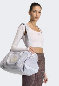 Vaaleanharmaa matkakassi verkkokoristein, kaksoiskahvoin ja säädettävällä olkahihnalla. Sileäpintainen, sisäosassa säilytystasku.