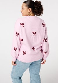 Vrouw in een lichtroze trui met donkerrode strikpatronen, gecombineerd met lichtblauwe jeans, die zijwaarts staat tegen een effen achtergrond.