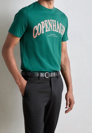 Mężczyzna w zielonej koszulce z napisem „COPENHAGEN” oraz czarnych spodniach z czarnym paskiem, stojący z jedną ręką w kieszeni.