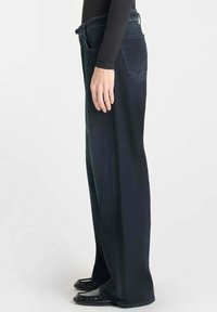 Jeans en denim foncé à jambes larges avec une texture lisse, taille mi-haute et détails sur les poches arrière. Modèle portant des ballerines noires.