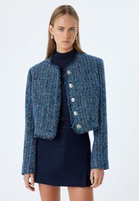 Blå tweedjakke med frynsekant og fem sølvknapper, parret med en mørkeblå struktureret kjole. Lange ærmer, cropped snit.