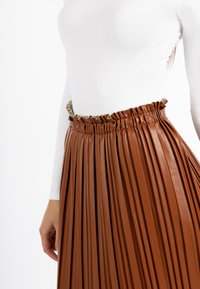 Bruine geplooide faux leren rok met een elastische tailleband en een gerimpelde groene accent. Draagt over een aansluitend wit langmouwtop.