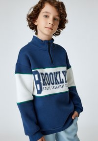 Sudadera con cremallera en azul marino y blanco con el texto "BROOKLYN". Presenta detalles en verde y un cuello alto. Fabricada con material suave y texturizado.