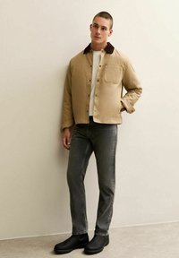 Ung man som bär en beige jacka med mörkrandig krage, vit skjorta, grå jeans och svarta kängor, står mot en ljus vägg.