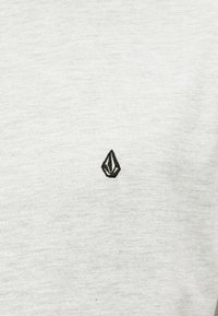 T-shirt gris avec une texture lisse comportant un petit logo noir brodé sur le côté gauche, ressemblant à une forme de diamant.