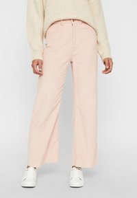 En person som bär en krämfärgad stickad tröja och ljusrosa vida byxor, i kombination med vita sneakers, står framför en neutral bakgrund.