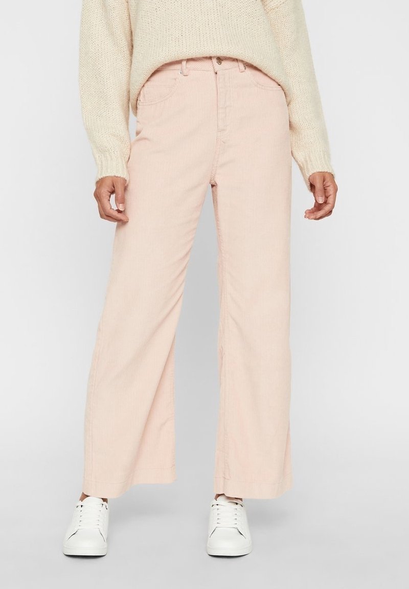 En person som bär en krämfärgad stickad tröja och ljusrosa vida byxor, i kombination med vita sneakers, står framför en neutral bakgrund.