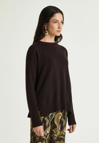 Maglione marrone lavorato a maglia con scollo tondo, spalle morbide e bordi a coste. Abbinato a pantaloni fantasia con disegni floreali.