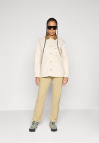 Persoon met lange vlechten die een oversized beige jack, tan broek, grijze wandelschoenen en grote donkere zonnebril draagt, staand tegen een witte muur.